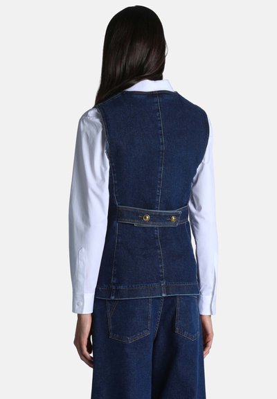 Personne portant un gilet en jean bleu foncé avec une bride arrière boutonnée par-dessus une chemise blanche à manches longues, tourné dos à la caméra.