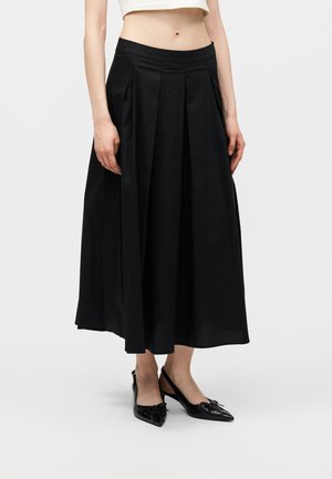 Frau trägt einen schwarzen, hoch taillierten, plissierten Midirock, kombiniert mit schwarzen spitzen Slingback-Ballerinas, steht vor einem schlichten Hintergrund.