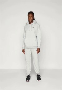 Gray hoodie and sweatpants set in soft fabric, featuring a logo on the chest and side. Hood draws with adjustable cords. Black athletic shoes.