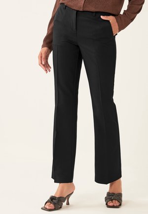 Femme portant un pantalon noir ajusté, un haut marron à manches longues et des sandales noires à talons ouvertes, avec une main dans la poche.