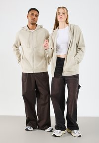 Beige mikina na zip s kapucí a šňůrkami, v kombinaci s volnými černými kalhotami. Obojí doplněno sportovními teniskami. Neutrální pozadí s šedou podlahou.