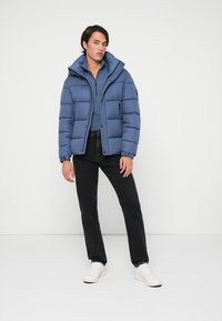 Blauer Steppjacke mit hohem Kragen, Reißverschluss vorne und gerippten Bündchen. Dazu ein blauer Langarm-Oberteil und schwarze Jeans. Weiße Sneakers.