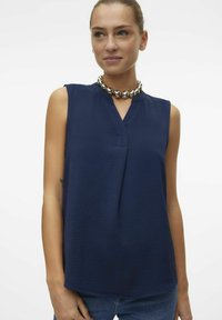 Blouse sans manches bleu marine avec un col en V et un tissu texturé. Le modèle porte un collier en argent chunky. L'arrière-plan est blanc uni.