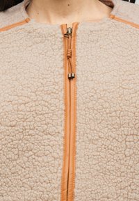 Giacca in pile beige con superficie strutturata, dotata di chiusura frontale con zip color cuoio e dettagli in pelle intorno al collo e alla zip.
