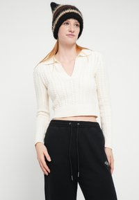 Krem pulover z vzorcem kit in V-izrezom ter dolgimi rokavi, kombiniran s črnimi trenirkami z elastičnim pasom in izvezenim logotipom. Pletena črtasta kapa.