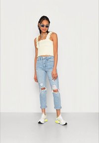 Top curto de cor clara, com nervuras e alças com frunces, combinado com jeans claros de cintura alta e destroçados, além de ténis brancos com detalhes em néon.