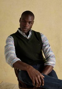 Sedíci muž s tmavou pokožkou, ktorý má na sebe čierny sveter bez rukávov cez viacfarebnú kockovanú košeľu a tmavé džínsy, pred béžovou stenou.