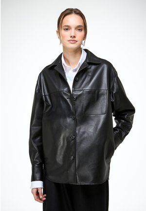 Young woman wearing a black leather button-up shirt over a white collared shirt, standing against a plain light background.