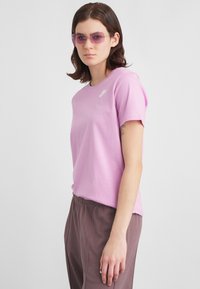 Νεαρή γυναίκα με γυαλιά πορφυρού τόνου, ελαφριά ροζ μπλούζα Nike και φαρδιά καφέ παντελόνια, που στέκεται μπροστά σε απλό φωτεινό φόντο.