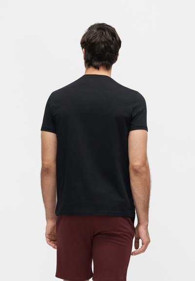 Άνδρας με κοντά σκούρα μαλλιά που φοράει μονόχρωμο μαύρο κοντομάνικο μπλουζάκι και μπορντό σορτς, στέκεται με την πλάτη σε ουδέτερο φόντο.