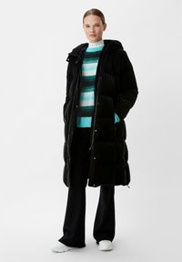 Black puffer coat with a hood, featuring a quilted design, zippered front, and side pockets. Worn over a green striped sweater and black pants.