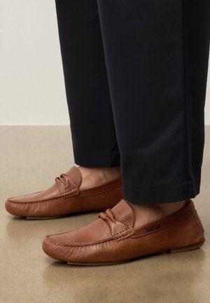 Bruine leren instap loafers gedragen met zwarte cropped broek op een beige vloer.
