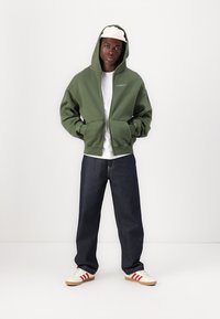 Grüne Zip-Hoodie mit Kapuze und Kordelzug, getragen über einem weißen T-Shirt, kombiniert mit dunkelblauen Jeans und cremefarbenen Sneakern mit roten Streifen.