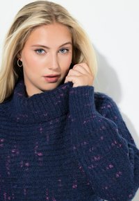 Granatowy, dzianinowy sweter z wysokim kołnierzem, z teksturowym wzorem i różowymi plamkami. Model trzyma kołnierz, eksponując szczegóły.