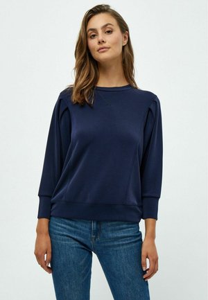 Felpa blu navy con maniche lunghe, collo a giro e polsini a coste. Presenta pieghe sottili sulle spalle e una texture del tessuto liscia.