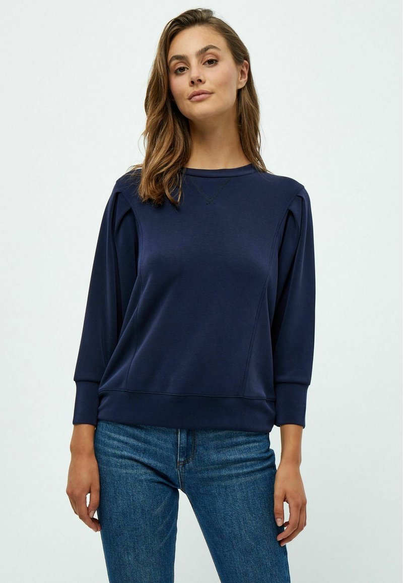 Felpa blu navy con maniche lunghe, collo a giro e polsini a coste. Presenta pieghe sottili sulle spalle e una texture del tessuto liscia.
