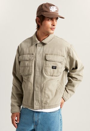 Beige spijkerjasje met een kraag, twee opgezette zakken aan de voorkant en een merklabel. De stof lijkt stevig en heeft gestikte details.