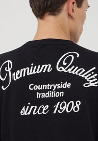 T-shirt in cotone nero con testo bianco sul retro: "Qualità Premium," "Tradizione rurale," e "dal 1908," in diversi caratteri.
