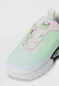 Baltas sporta apavi ar neona zaļiem akcentiem, sieta augšdaļa, apļveida šņores un "Air Max" logo uz mēles cilpas.