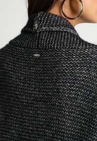 Frau, die einen grob gestrickten, dunkelgrauen Pullover mit einem kleinen metallischen "Guess"-Logo auf dem oberen Rücken und großen goldenen Creolen trägt.
