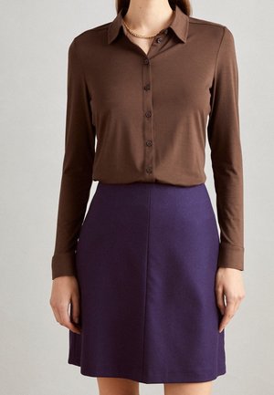 Femme portant une chemise marron à manches longues boutonnée, rentrée dans une jupe violette taille haute, avec un collier chaîne en or visible.