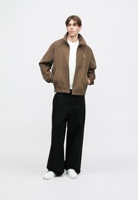 Brun semsket bomberjakke med ribbede mansjetter, glidelås og en liten logo, paret med svarte vide bukser og hvite sneakers.