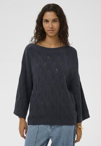 Marineblauer Strickpullover mit wellenförmigem Muster, weitem Ausschnitt und dreiviertellangen Ärmeln, kombiniert mit hellblauen Jeans.
