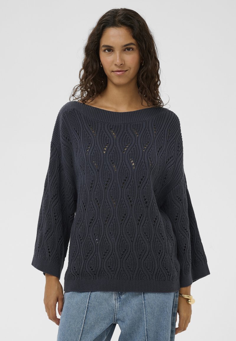 Marineblauer Strickpullover mit wellenförmigem Muster, weitem Ausschnitt und dreiviertellangen Ärmeln, kombiniert mit hellblauen Jeans.