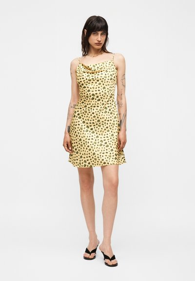 Γυναίκα με σκούρα μαλλιά και τατουάζ που φοράει κίτρινο φόρεμα με μαύρες βούλες και μαύρα σανδάλια με τακούνι σε λευκό φόντο.