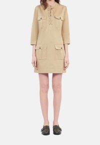 Woman wearing a beige knee-length dress with four buttoned pockets and black slip-on shoes, standing against a plain light background.