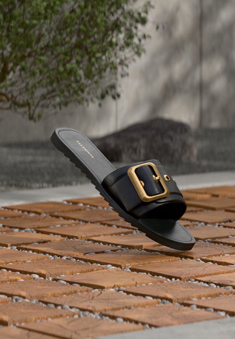 Μαύρο σανδάλι με φαρδιά λουρί και μεγάλη χρυσή αγκράφα, τοποθετημένο σε ξύλινο δάπεδο με πράσινο φυτό και γκρι τοίχο στο φόντο.