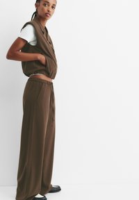 Top marrone a coste, senza maniche, con scollatura rotonda, abbinato a pantaloni con cordoncino coordinati. Il modello indossa scarpe nere, con una maglietta chiara all'interno.
