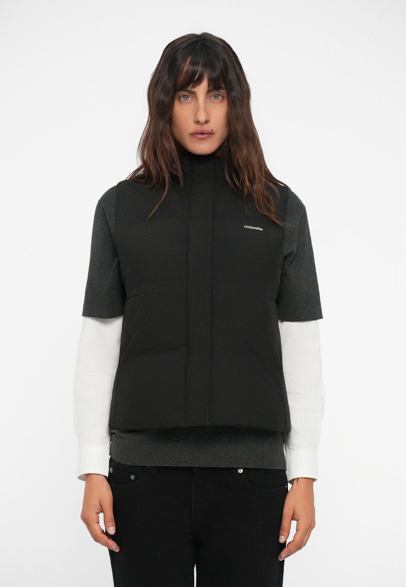 Gilet imbottito nero senza maniche con colletto alto, tasche frontali e logo. Indossato sopra una maglietta corta grigia e una camicia a maniche lunghe bianca.
