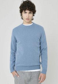 Pull en tricot bleu clair avec un col rond, des poignets et un ourlet côtelés. Porté sur une chemise blanche, présentant une texture lisse.