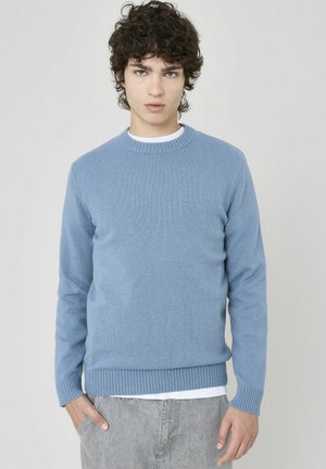 Pull en tricot bleu clair avec un col rond, des poignets et un ourlet côtelés. Porté sur une chemise blanche, présentant une texture lisse.