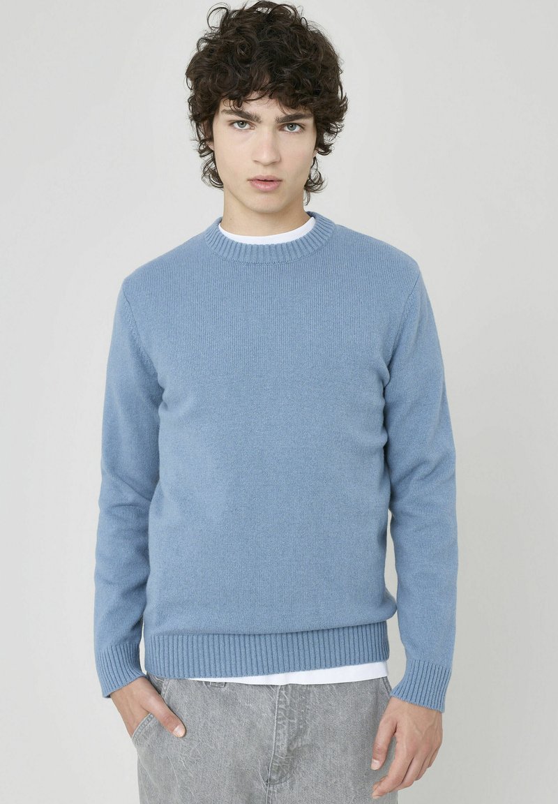 Pull en tricot bleu clair avec un col rond, des poignets et un ourlet côtelés. Porté sur une chemise blanche, présentant une texture lisse.