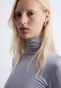 Grauer Rollkragenpullover mit langen Ärmeln und glatter Textur. Das Modell hat lange, glatte blonde Haare und kleine silberne Creolen.