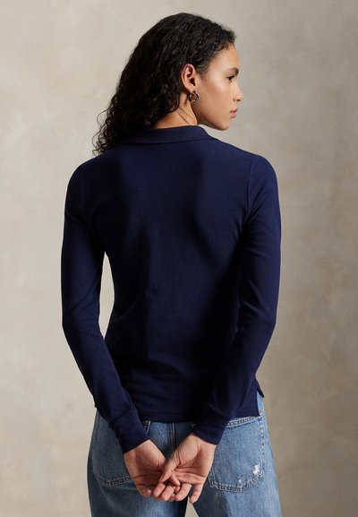 Tamsiai mėlynas ilgomis rankovėmis megztinis su polo apykakle, pagamintas iš minkštos medžiagos, su šoninėmis plyšiais ir prigludusia forma. Dėvimas su šviesiai mėlynomis džinsais.