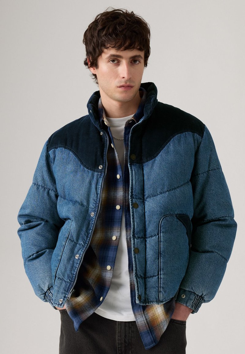 Blå denim pufferjakke med højt krave, kordfløjsdetaljer på skuldrene og elastiske manchetter. Under jakken ses en ternet flannel skjorte.
