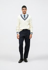 Balta megzta megztinio megztinė su tamsiai mėlynomis detalėmis, šviesiai mėlyna suknelės marškiniai, tamsiai mėlynos kelnės ir juodi batai. Megztinis turi V formos iškirptę.