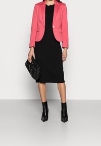 Rosa tætsiddende blazer over en sort ribbet strik kjole, parret med en sort quilted håndtaske og spidse sorte ankelsko.
