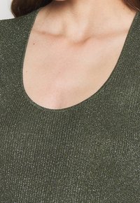 Haut vert olive avec un col en V profond, fabriqué en tissu côtelé avec une texture subtilement scintillante. Aucune décoration visible.