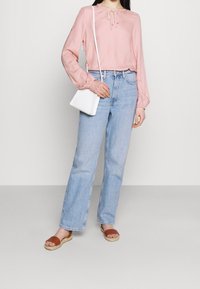 Blusa rosa chiaro a maniche lunghe con dettaglio a fiocco, jeans chiari a vita alta, sandali piatti marroni e una piccola borsa a tracolla bianca.