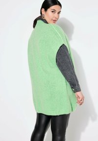 Ljusgrön oversized sweatshirtväst i fluffig stickad kvalitet. Har vida ärmhål och ribbat nederkant. Bärs över en mörk denimskjorta.