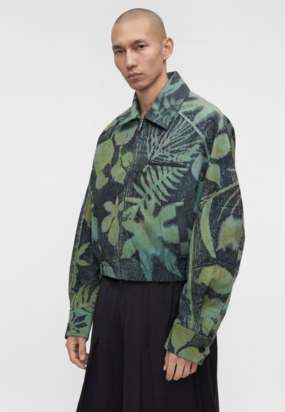 Άνδρας με ξυρισμένο κεφάλι που φοράει πράσινο και μαύρο μπουφάν με φερμουάρ και βοτανικό σχέδιο και μαύρο πιετέ φαρδύ παντελόνι, στέκεται μπροστά σε μονόχρωμο φόντο.