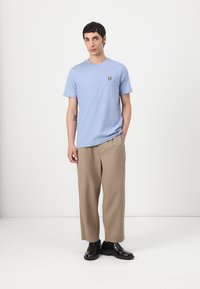 T-shirt bleu clair à manches courtes avec une finition texturée et un petit logo jaune sur la poitrine, associé à un pantalon beige et des chaussures noires.