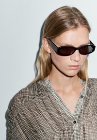 Žena s rovnými blond vlasy, oblečená v úzkých černých slunečních brýlích a knoflíkované, světle hnědé kostkované blůze na jednobarevném světlém pozadí.