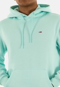 Sudadera verde menta claro hecha de una tela suave, con capucha ajustable, bolso frontal y un pequeño logo en rojo, blanco y azul.