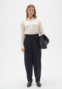 Cremefarbendes Satin-Hemd mit langen Ärmeln, schwarze weite Hose und schwarze Slipper. Modell hält eine schwarze Ledertasche.