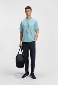 Camiseta azul claro con un patrón gráfico en gris y negro, combinada con pantalones oscuros y una bolsa oscura. La tela parece suave y casual.